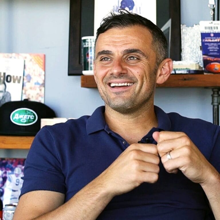 Ein lächelnder Mann mit kurzen dunklen Haaren und Bartstoppeln, der ein marineblaues Poloshirt trägt, sitzt in einem Raum. Hinter ihm stehen Regale mit Büchern, einem Hut, gerahmten Fotos und verschiedenen Gegenständen, die eine lockere, persönliche Büroatmosphäre schaffen.