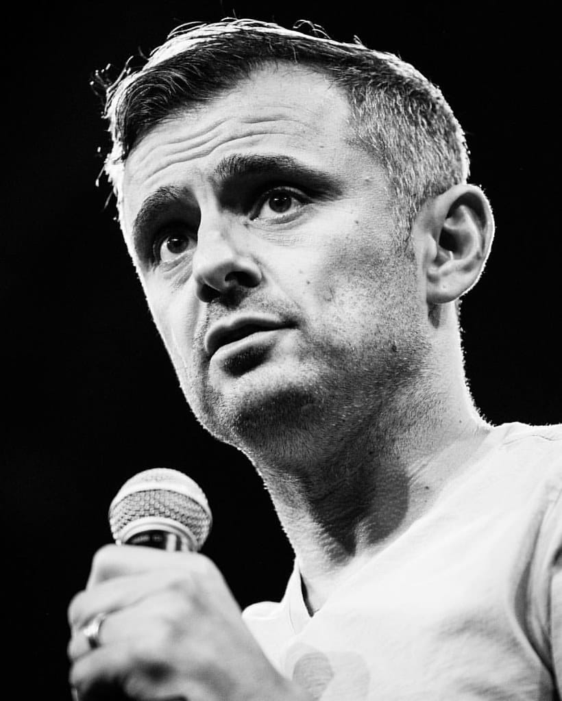 Ein Mann mit kurzen Haaren und Bartstoppeln hält ein Mikrofon und spricht aufmerksam. Er trägt ein helles Hemd und blickt leicht nach oben. Das Foto ist schwarz-weiß, mit dramatischer Beleuchtung, die seine Gesichtszüge vor einem dunklen Hintergrund hervorhebt.